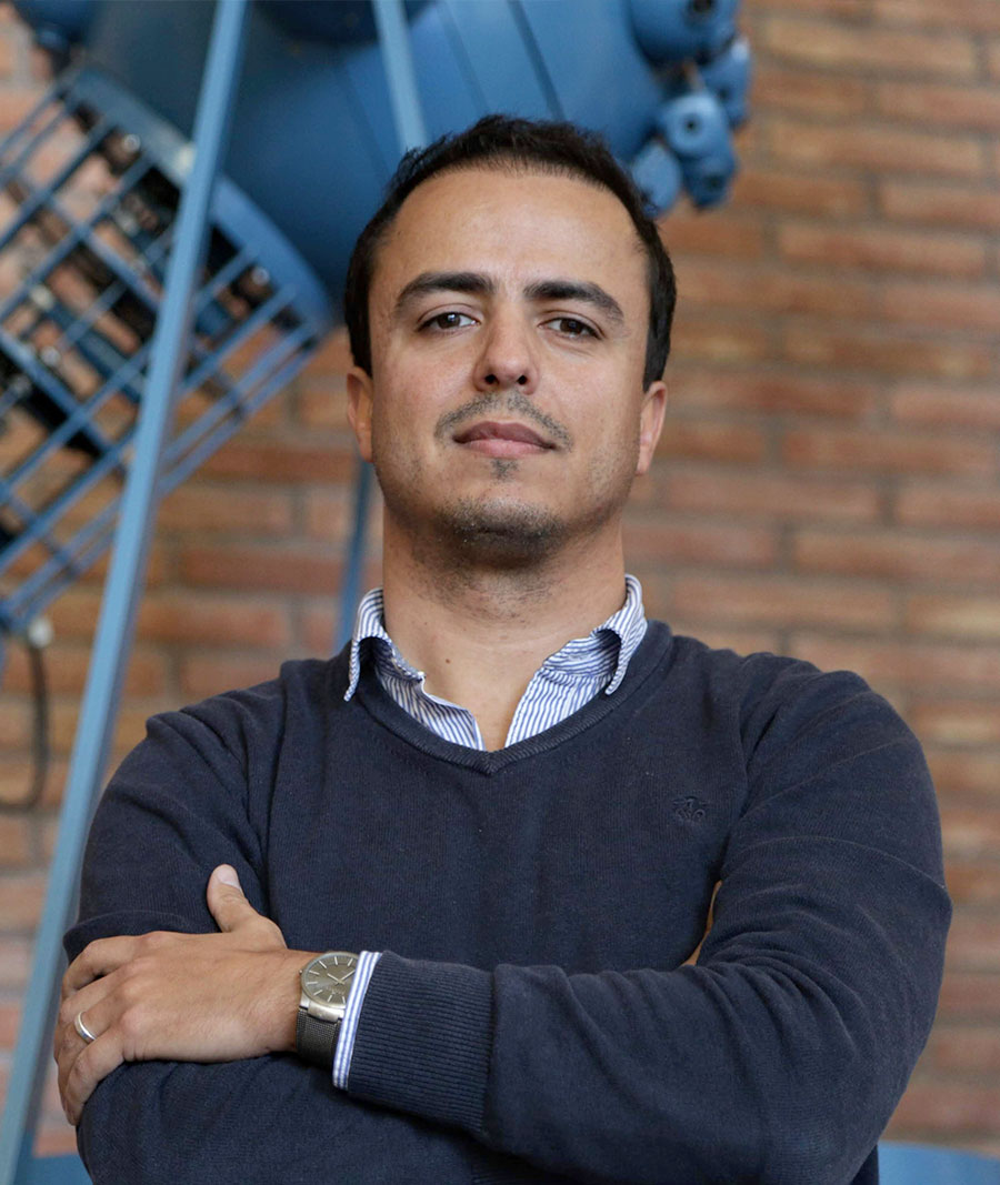 A person dressed in a dark blue jumper and striped shirt poses for a photo with their arms crossed. There is a large blue machine and a red brick wall in the background.